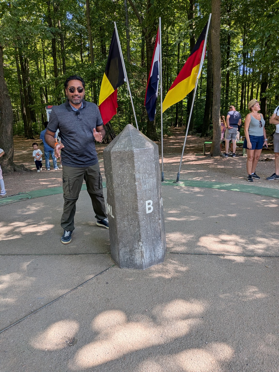 Saki The Wanderer standing beside the Drielandenpunt tripoint stone marked B and NL, with the flags of Belgium, the Netherlands and Germany behind it