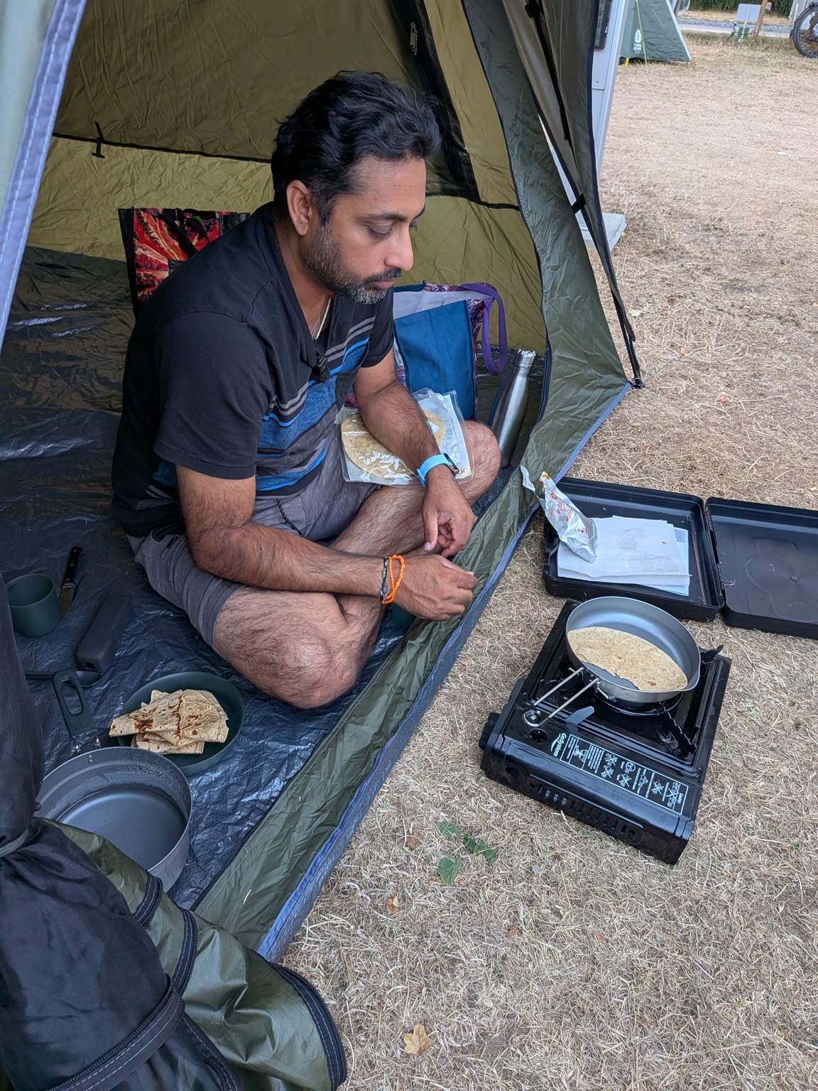 Saki The Wanderer sitting in a tent cooking rotis on a portable gas stove, with a packet of ready-made Dal Makhani nearby