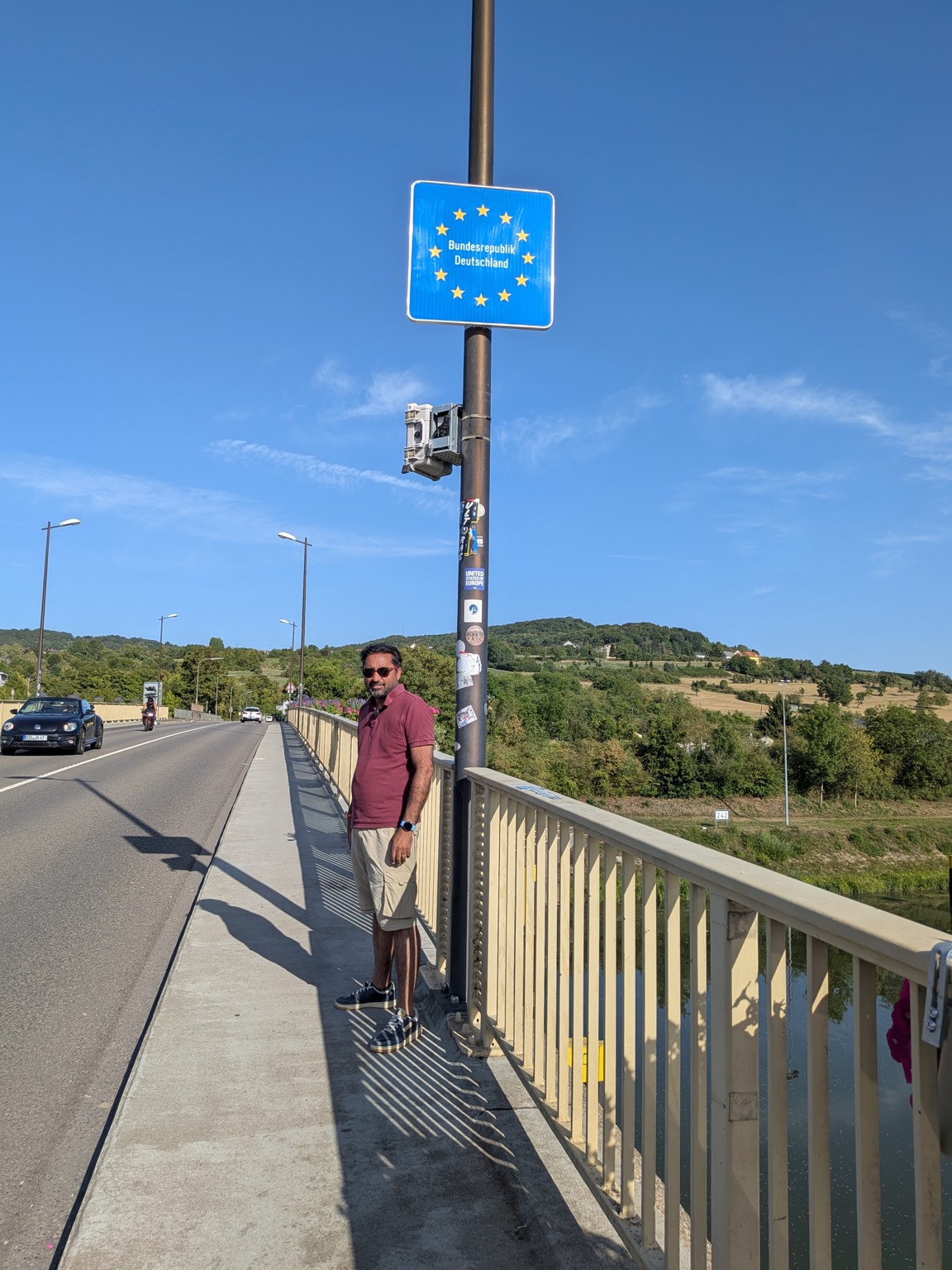 Saki The Wanderer standing on a bridge next to a blue EU-style sign reading Bundesrepublik Deutschland, marking the border into Germany near Schengen